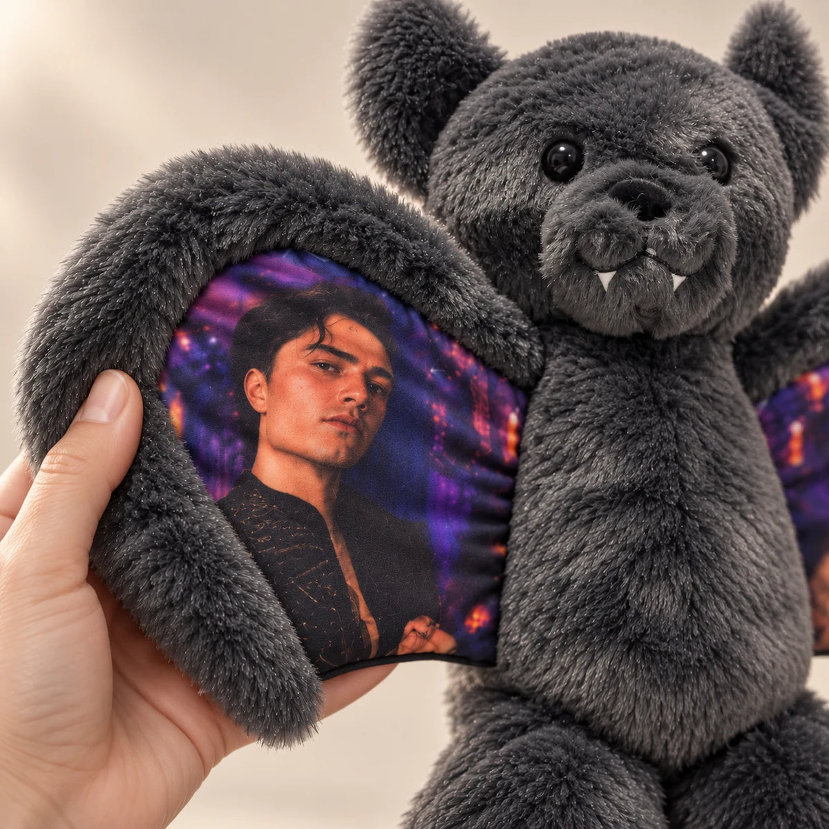 Close-up of a hand holding a gray bat bear's plush wing with a portrait printed on the fabric.